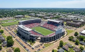 Aerial view of a large stadium filled with red-clad spectators, surrounded by parking lots, buildings, and trees.