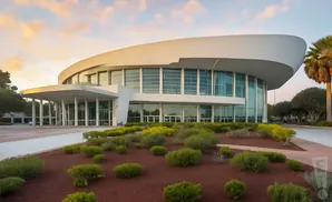 Modern, circular white building with large windows and an entrance canopy, surrounded by landscaping and palm trees at sunset.