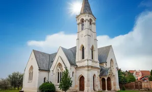 Exterior of a pale stone church with a tall steeple, under a bright blue sky with the sun shining.