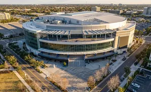 Aerial view of an outdoor sports arena with a large glass facade, surrounded by trees and parking lots.