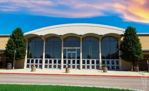 Modern building entrance with large glass doors and windows, arched canopy, and trees, under a colorful sky.