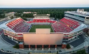 Aerial view of a large, modern football stadium with red seats, a green field, and surrounding trees.