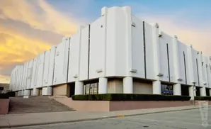 Modern white building with many vertical accents and rounded pillars, under a cloudy, colorful sky.