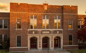 Exterior of an old brick school building with arched doorways and many windows reflecting the sunset.