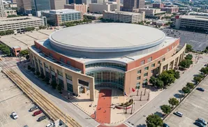 Aerial view of a large, circular arena with a gray roof and a beige and red exterior, surrounded by parking lots and city buildings.