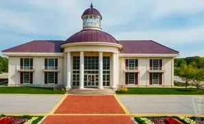 A large, tan building with a maroon domed roof and a cupola, fronted by white columns and glass doors, with red brick pathways and landscaping.
