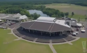 Aerial view of a large outdoor amphitheater with a dark roof and tiered seating, surrounded by trees and a small lake.