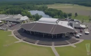 Aerial view of a large amphitheater with tiered seating facing a covered stage, surrounded by buildings and a parking lot.
