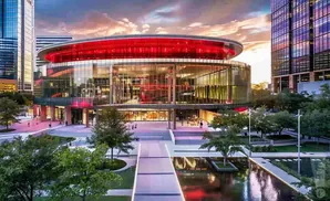 A modern, circular glass building with a red roof, surrounded by trees and reflecting pools, at sunset.