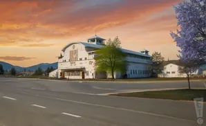 A white barn-like building with a curved roof stands under a vibrant orange sunset sky, with a road in the foreground.