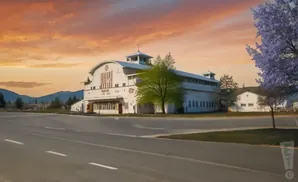 A white barn with a curved roof stands beside a road under a colorful sunset sky.