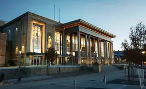 Grand stone building with large windows and columns lit up at dusk, with trees and streetlights outside.
