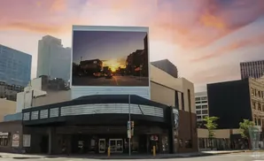 A marquee-style theater in a city is covered by a large digital billboard showing a sunset over a street.