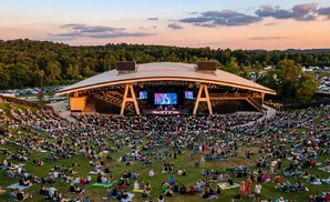 Outdoor concert with a packed lawn seating area and a large stage under a curved roof.