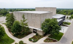 Aerial view of a large concrete building with modern dark gray additions and surrounding trees and parking lots.