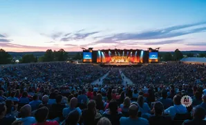 Outdoor concert at dusk with a large audience watching a band perform on a brightly lit stage.