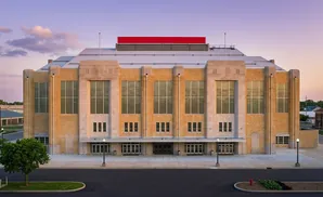 The Coliseum, a large art deco building with yellow brick and stone accents, stands under a soft purple sunset.