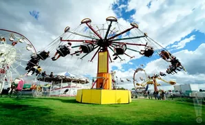 A swing ride spins with people at a fair, with a Ferris wheel and carousel in the background.