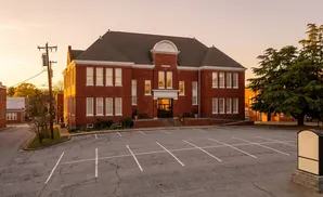 Sunset over a red brick building with a parking lot in front and large evergreen trees to the right.