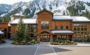 A modern wooden lodge with large windows sits at the base of snow-covered mountains and pine trees.