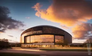Modern arena with glass facade at sunset, reflecting warm orange clouds in the sky.