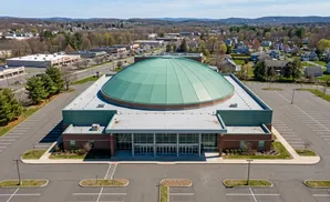 An aerial view of a large building with a green domed roof and a brick facade, surrounded by parking lots and greenery.