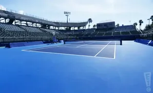 A bright blue tennis court with stadium seating and palm trees under a clear sky.