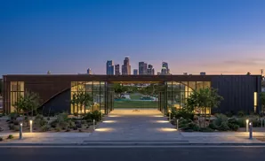 Modern building with glass walls and wooden louvers opens to a park with a city skyline at dusk.
