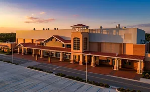Aerial view of a large building with a clock tower at sunset, featuring a red roof and beige walls, set against a colorful sky.