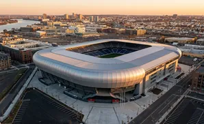 Drone view of a modern soccer stadium at sunset, with a cityscape and river in the background.