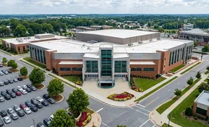 Aerial view of a large brick and glass building with a white roof, surrounded by trees and a parking lot.