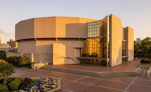 Modern concrete building with glass facade, tiered plaza, and flowerbeds in warm sunset light.