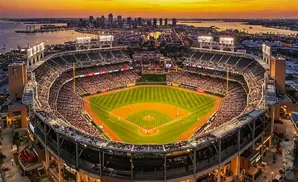 Aerial view of a baseball stadium at sunset with a city skyline and bay in the background.