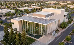 Aerial view of a modern building with solar panels and glass facade, surrounded by trees and parking lots.