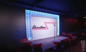 A comedy club stage with a cityscape backdrop, a microphone, and red-draped tables.