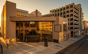 Modern building with large glass windows reflecting sunlight and nearby buildings, next to a multi-story parking garage.