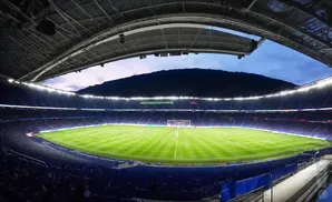 Wide angle view of a soccer stadium with a lit green field and blue seats, under a dusky sky.