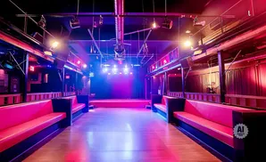 An empty nightclub with a stage, dance floor, and red velvet seating, lit by colorful stage lights.