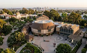 Aerial view of The Old Globe theater and the surrounding gardens in Balboa Park, San Diego, with the city skyline in the background.