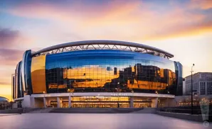 Modern stadium with reflective glass exterior, reflecting a colorful sunset sky and city skyline.