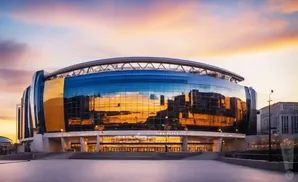 A modern stadium with a glass facade reflects a colorful sunset.