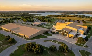 Aerial view of a large complex with multiple buildings, parking lots, and a body of water in the distance at sunset.