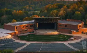 Outdoor amphitheater with a brick building, stage, and tiered seating in front of a wooded hillside.
