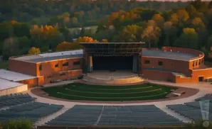 Outdoor amphitheater with a stage, grassy seating area, and rows of seats, framed by brick buildings and trees.