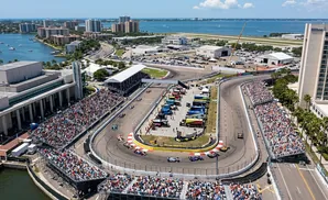 An aerial view of a race track with cars and spectators, set against a backdrop of city buildings and the ocean.