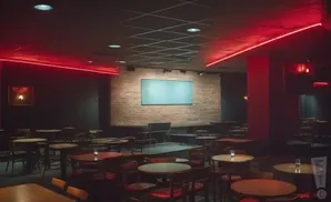 A dimly lit comedy club with red neon lights, brick wall stage, and tables and chairs.