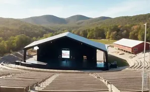 An outdoor amphitheater stage and seating area with a red barn in the background, set against a backdrop of tree-covered hills.