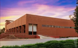 A modern brick building with a colonnade and a long horizontal window against a purple and orange sunset sky.