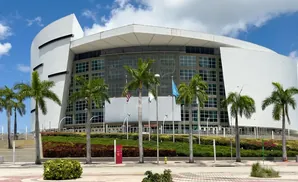 Modern white arena with glass facade, palm trees, and landscaped planters under a blue sky.