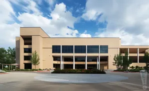 A modern, beige brick building with large windows and trees under a cloudy blue sky.